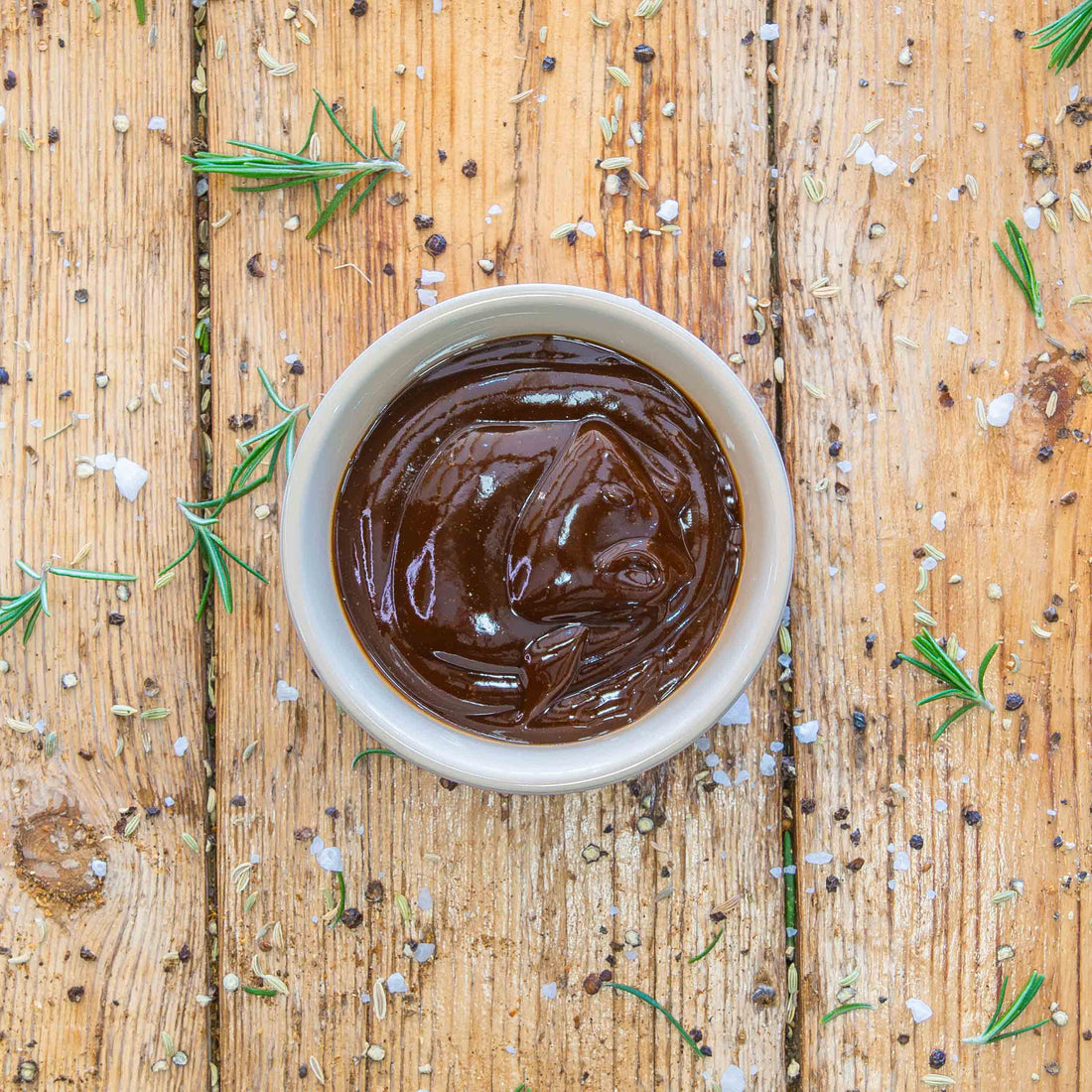 Padstal Deli branded smokey BBQ sauce in a bottle. A white ceramic bowl next to the bottle is filled with the brown colored sauce. Both are displayed on a vertically stacked light brown wooden panel backdrop, surrounded by cosmetic rosemary, salt and other spices