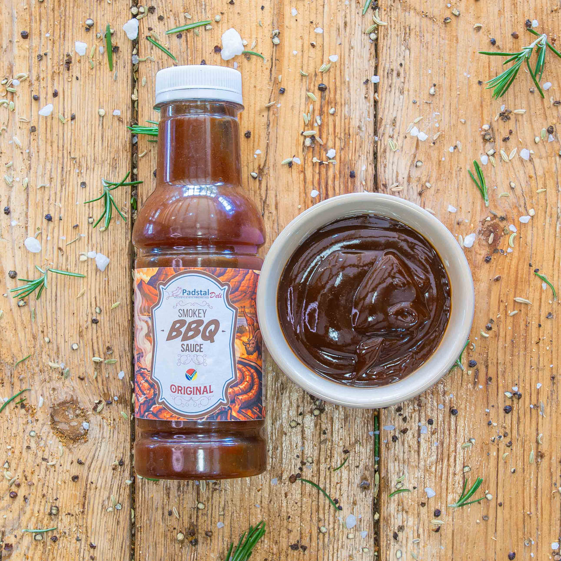 Padstal Deli branded smokey BBQ sauce in a bottle. A white ceramic bowl next to the bottle is filled with the brown colored sauce. Both are displayed on a vertically stacked light brown wooden panel backdrop, surrounded by cosmetic rosemary, salt and other spices