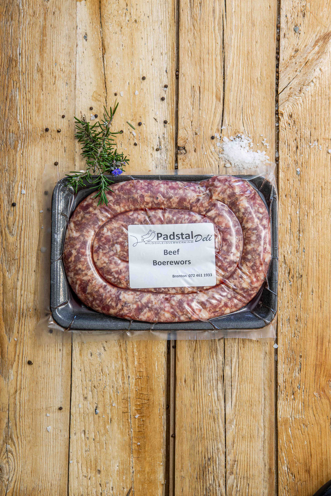 Padstal Deli Artisan Beef Boerewors coiled in a tight spiral on a light rustic wooden background, garnished with coarse sea salt, black peppercorns, and a fresh rosemary sprig.