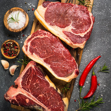 Padstal Deli premium raw beef collection feature, displaying thick-cut marbled steaks and a T-bone steak on a wooden board with coarse salt, peppercorns, and fresh red chilies.