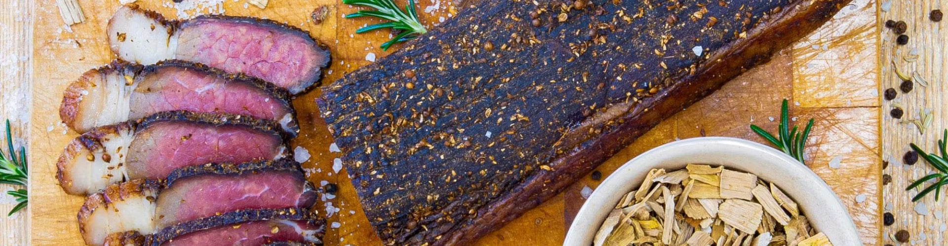 Premium South African beef biltong slab next to sliced biltong pieces on a wooden board, decorated with coriander, coarse salt, peppercorns, and fresh rosemary.