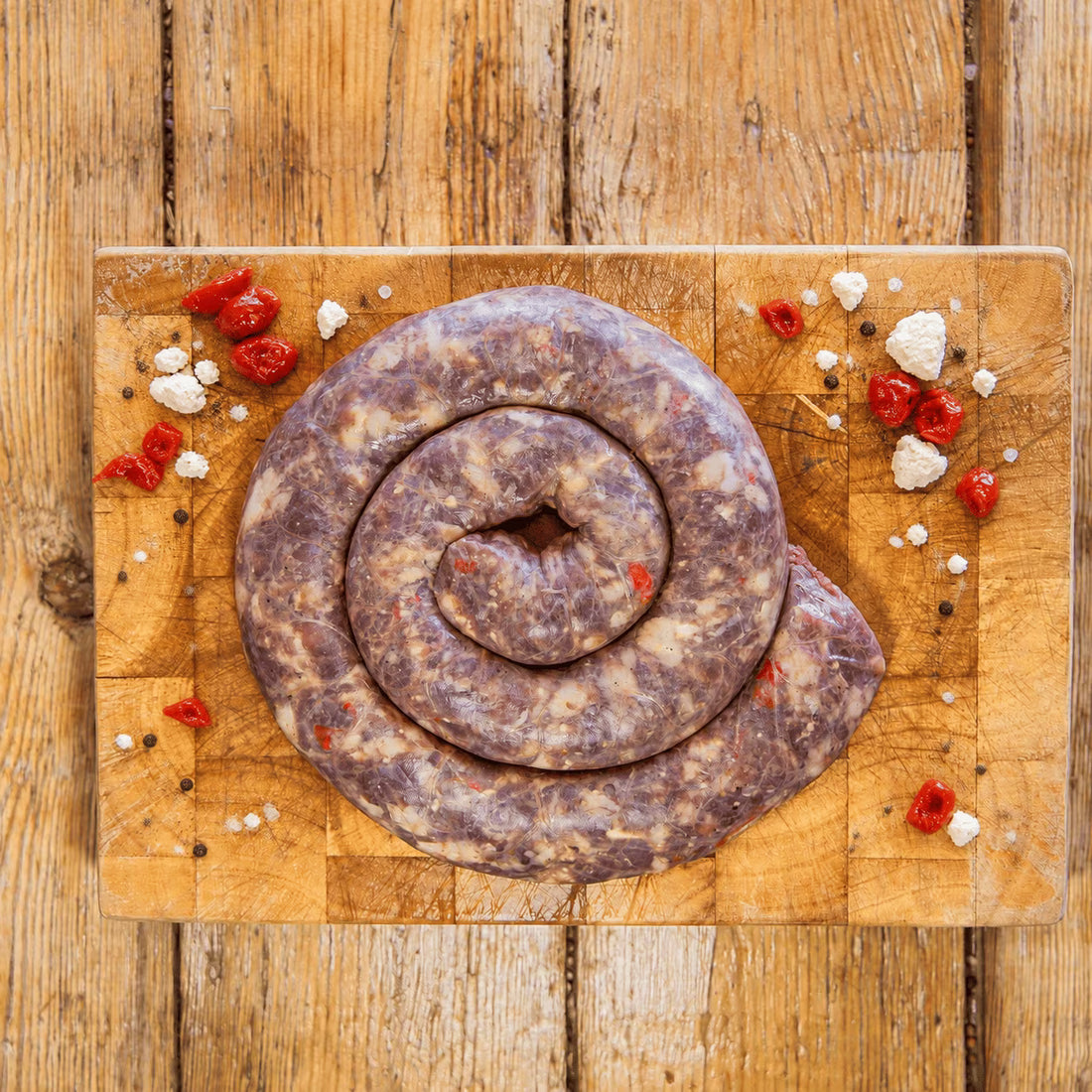 A raw, coiled boerewors sausage speckled with bits of red pepper and white cheese, resting on a well-used, square wooden chopping board. The board sits on a rustic wooden plank table garnished with scattered chopped red peppers, crumbled feta cheese, coarse salt, and whole black peppercorns.
