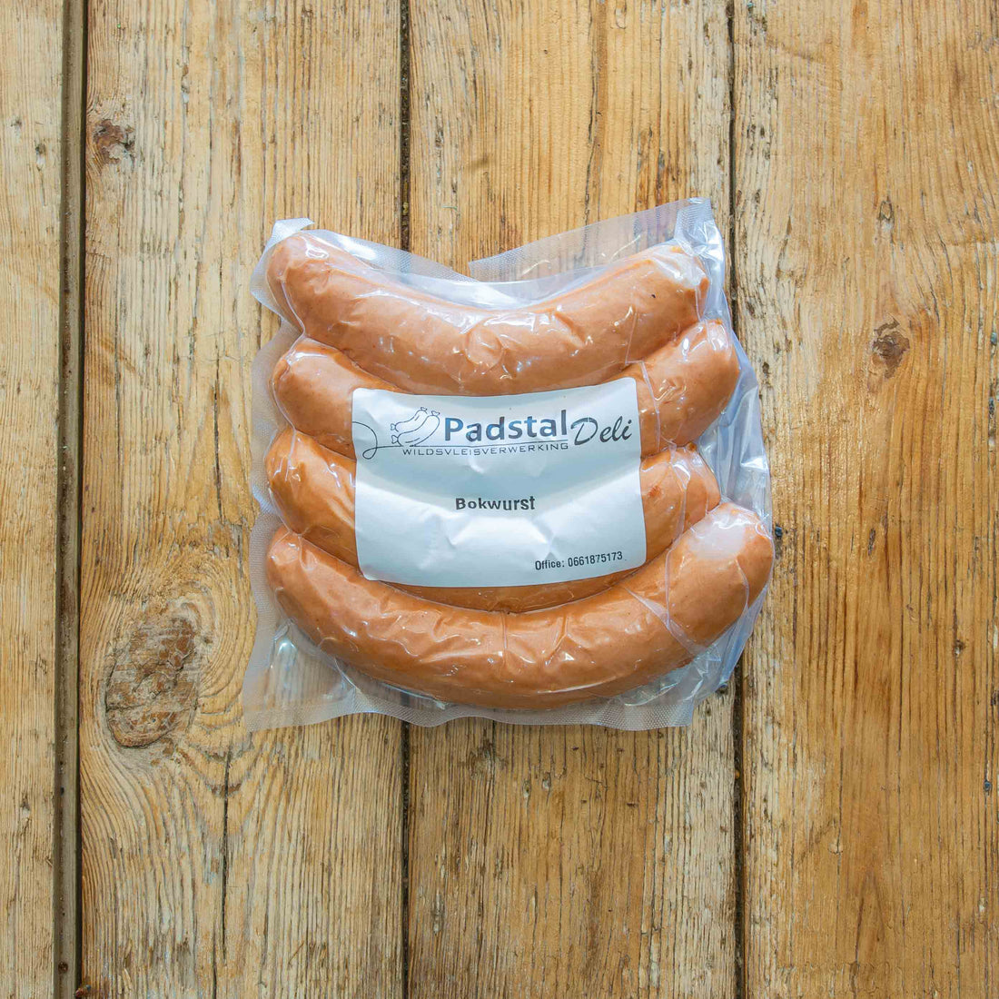 Padstal Deli Bokwurst set on a vertically stacked light wooden backdrop with decorative rosemary, salt and other spices around it.