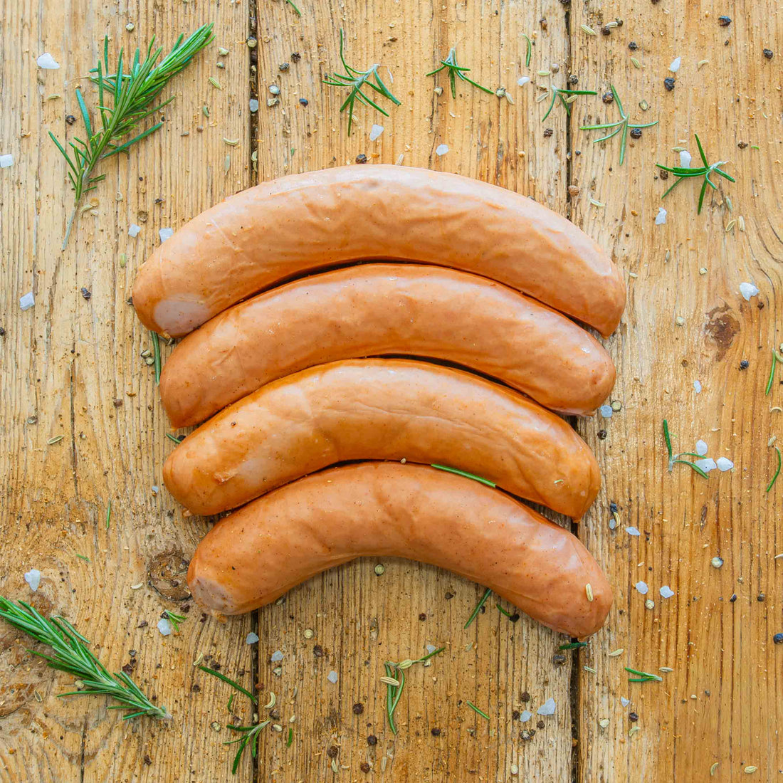 Padstal Deli Bokwurst set on a vertically stacked light wooden backdrop with decorative rosemary, salt and other spices around it.