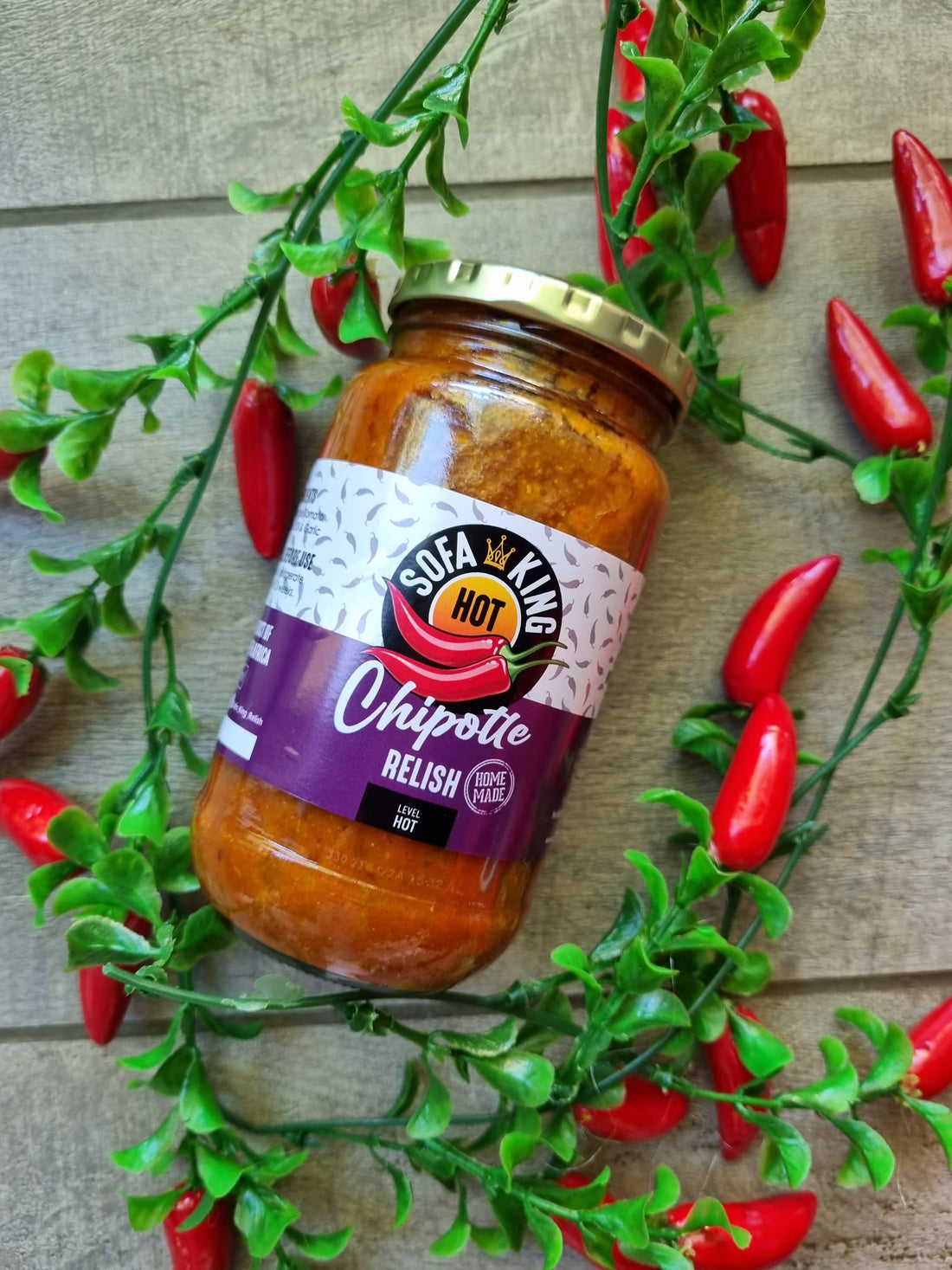 A glass jar of Sofa King Hot Chipotle Relish is displayed on a rustic wooden background, surrounded by decorative green foliage and red chili peppers.