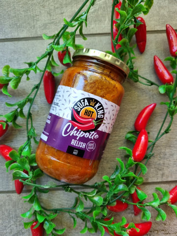 A glass jar of Sofa King Hot Chipotle Relish is displayed on a rustic wooden background, surrounded by decorative green foliage and red chili peppers.