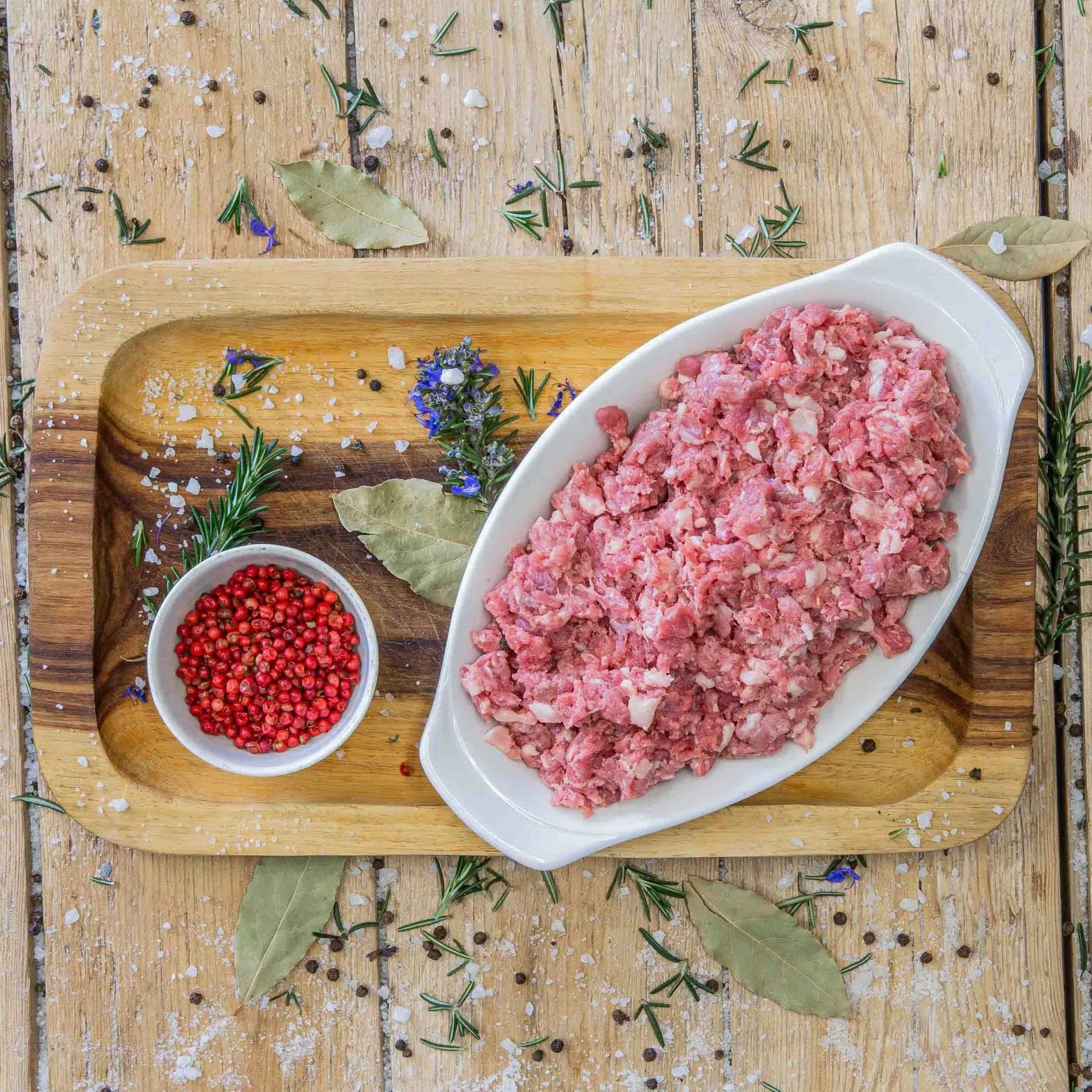 Padstal Deli premium raw Coarse Game Mince served in a white dish on a wooden platter, decorated with a small bowl of pink peppercorns, dried bay leaves, and fresh rosemary.
