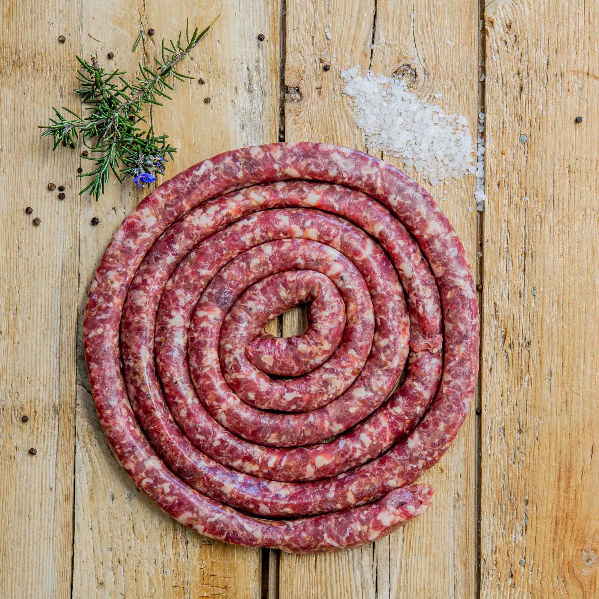 Padstal Deli Grabouw Braaiwors coiled in a tight spiral on a light wooden background, garnished with coarse sea salt, black peppercorns, and fresh rosemary.