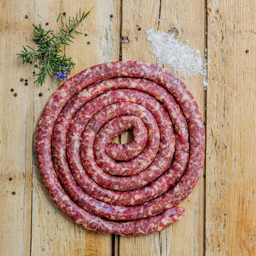Padstal Deli Grabouw Braaiwors coiled in a tight spiral on a light wooden background, garnished with coarse sea salt, black peppercorns, and fresh rosemary.