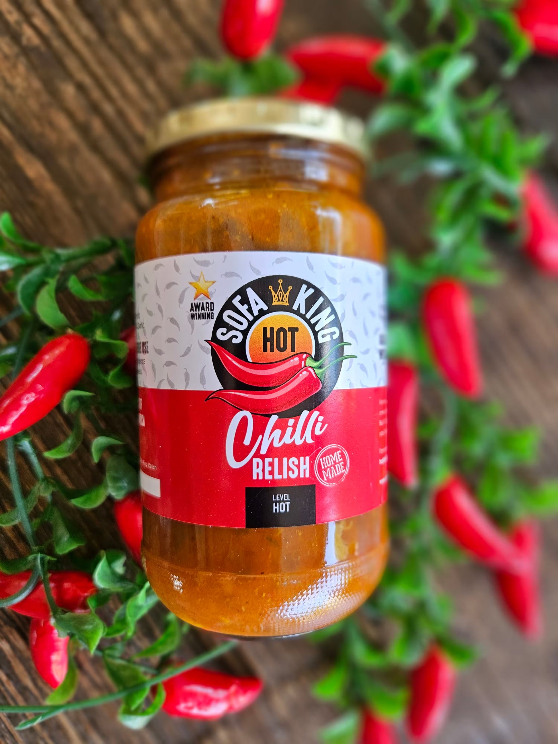 A glass jar of Sofa King Hot Chilli Relish is displayed on a rustic wooden background, surrounded by decorative green foliage and red chili peppers.