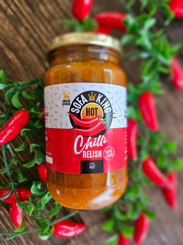 A glass jar of Sofa King Hot Chilli Relish is displayed on a rustic wooden background, surrounded by decorative green foliage and red chili peppers.