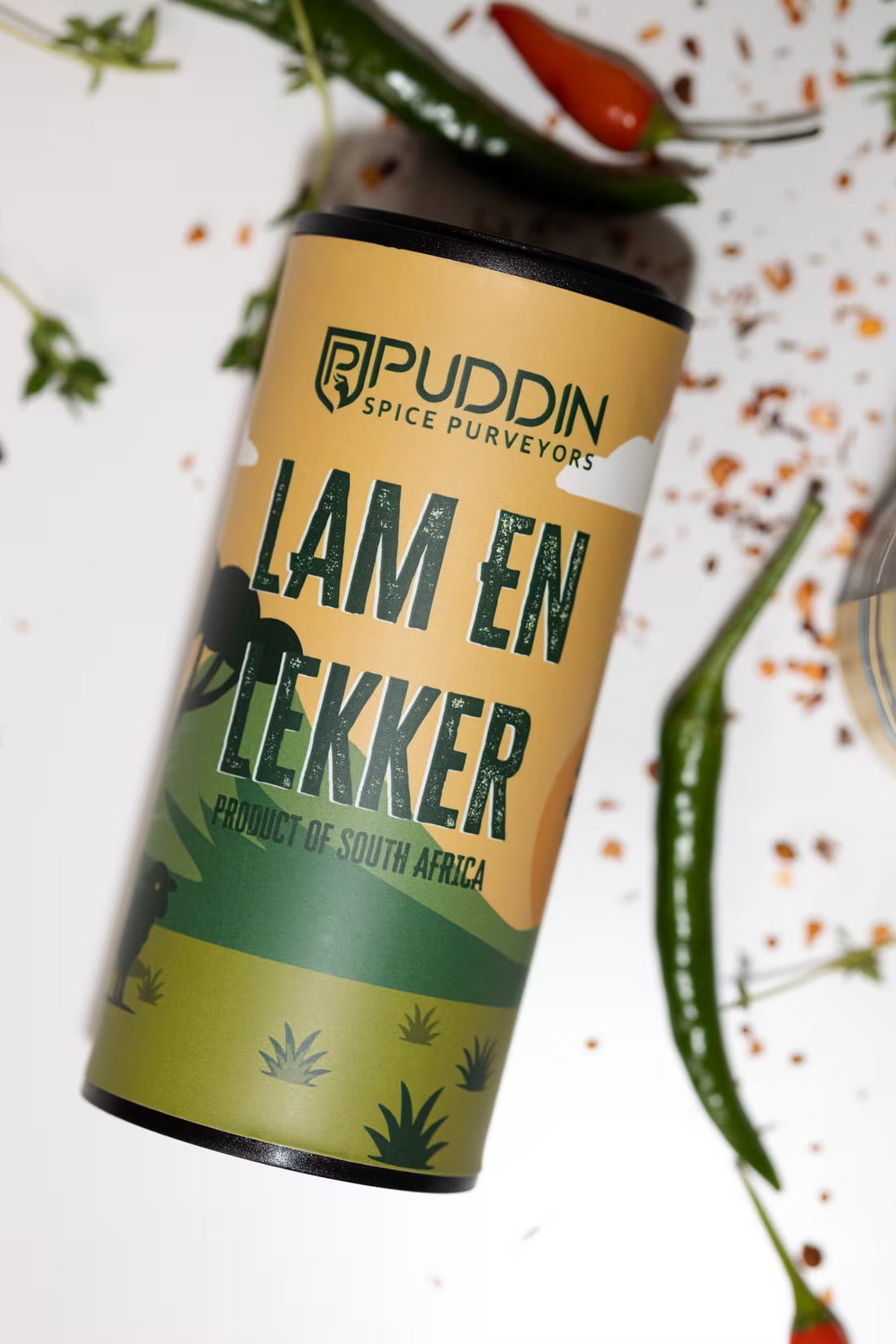 A cylindrical tin of Puddin Spice Purveyors 'Lam en Lekker' spice mix, styled diagonally on a white surface with scattered chili peppers and herbs.