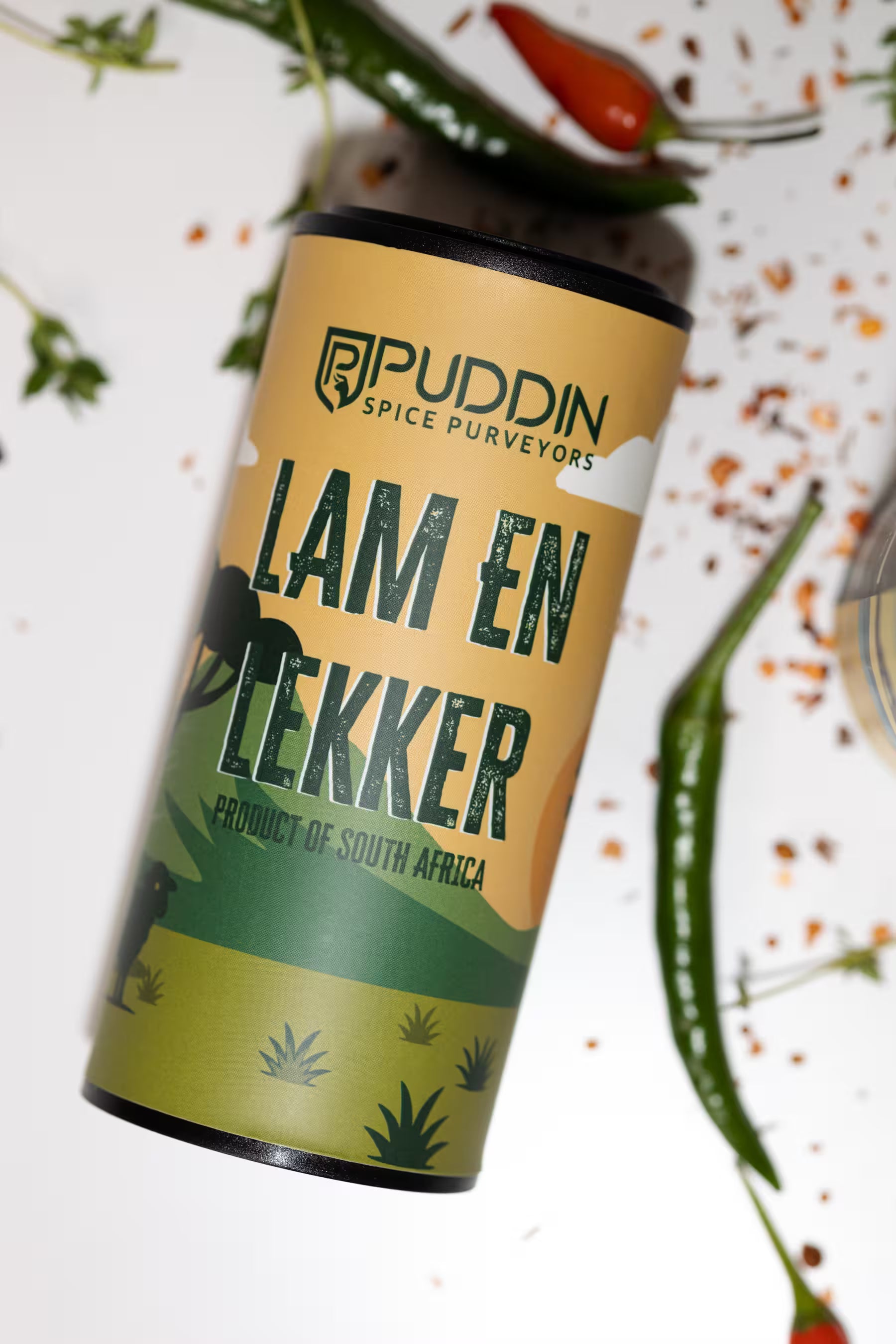 A cylindrical tin of Puddin Spice Purveyors 'Lam en Lekker' spice mix, styled diagonally on a white surface with scattered chili peppers and herbs.