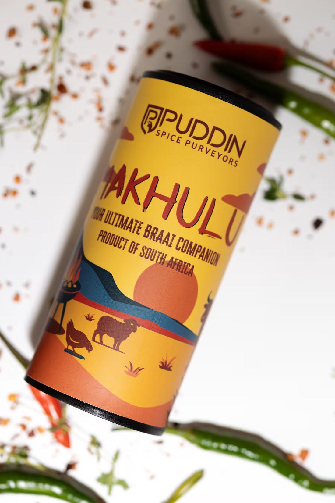 A cylindrical tin of Puddin Spice Purveyors 'Makhulu' spice blend, styled diagonally on a white surface with scattered red chili peppers and green chilies.