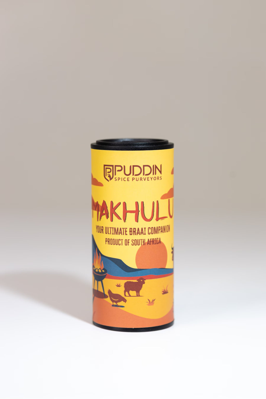A cylindrical tin of Puddin Spice Purveyors 'Makhulu' spice blend, styled diagonally on a white surface with scattered red chili peppers and green chilies.