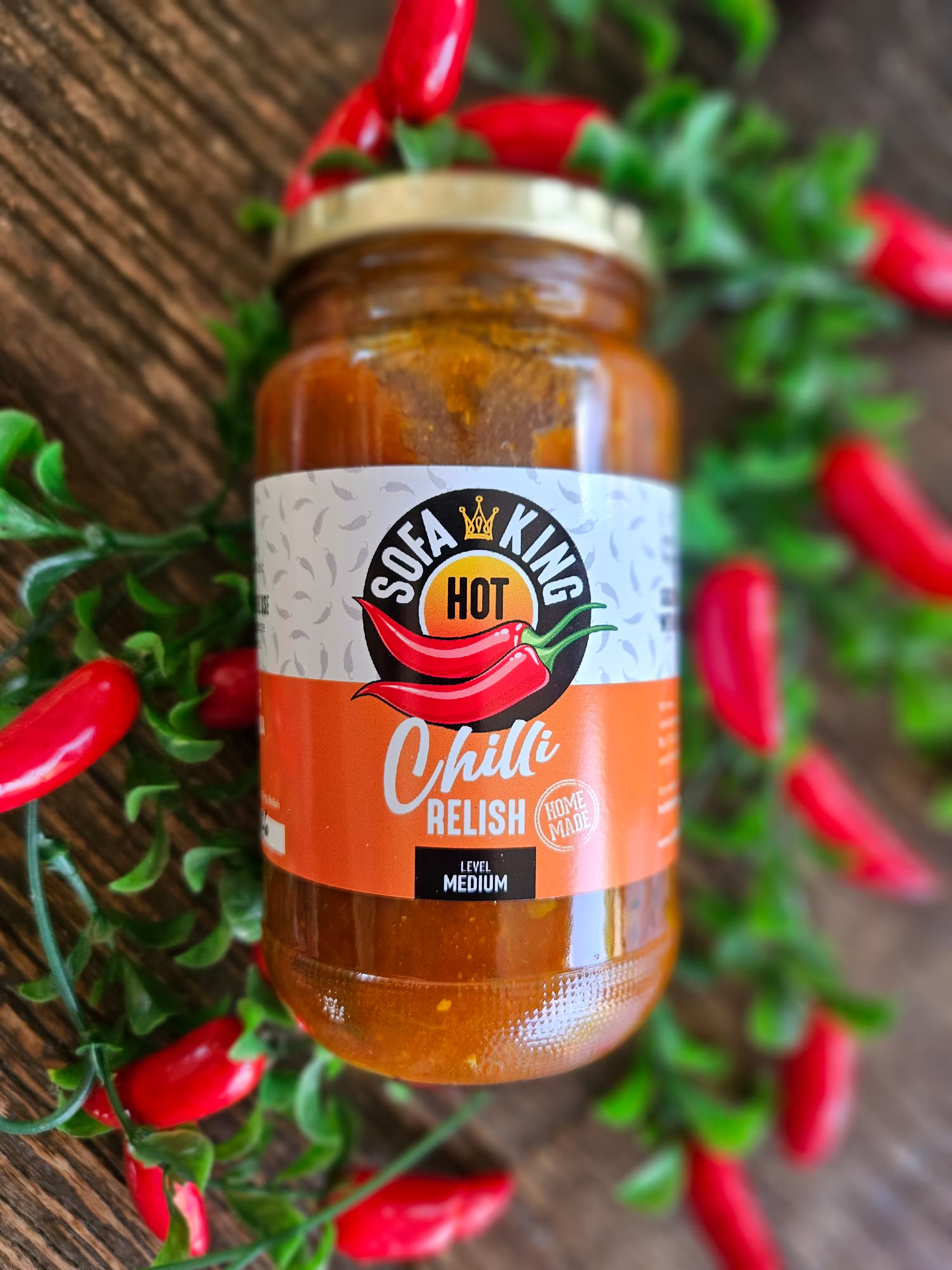 A glass jar of Sofa King Medium Chilli Relish is displayed on a rustic wooden background, surrounded by decorative green foliage and red chili peppers.