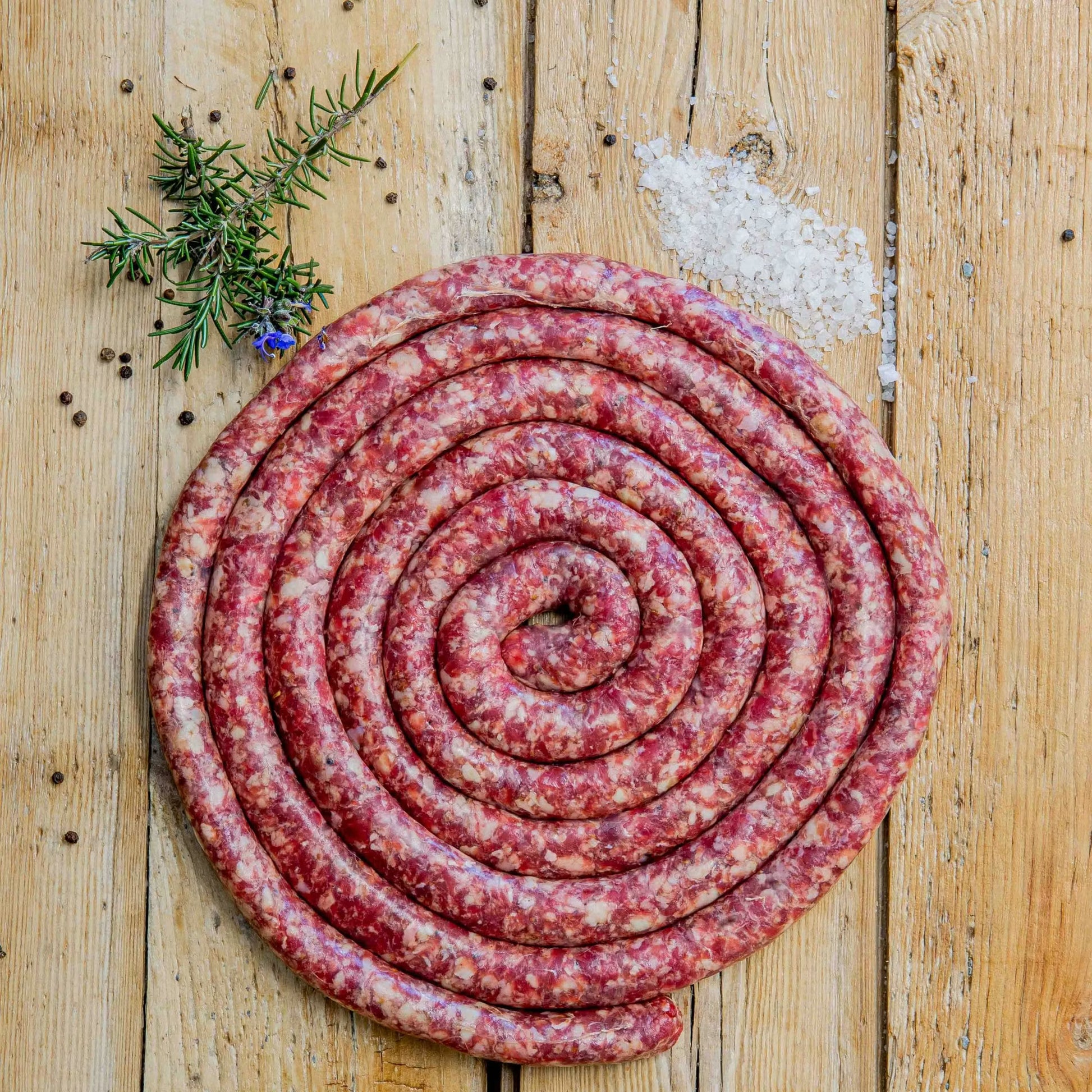 Padstal Deli Ontbyt Braaiwors coiled in a tight spiral on a light wood background, garnished with a pile of coarse sea salt, black peppercorns, and fresh rosemary.