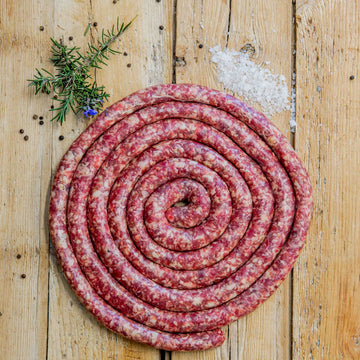 Padstal Deli Ontbyt Braaiwors coiled in a tight spiral on a light wood background, garnished with a pile of coarse sea salt, black peppercorns, and fresh rosemary.