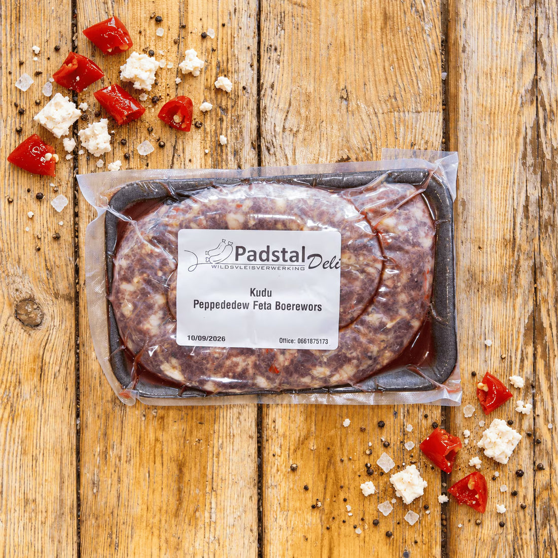 A raw, coiled boerewors sausage speckled with bits of red pepper and white cheese, resting on a well-used, square wooden chopping board. The board sits on a rustic wooden plank table garnished with scattered chopped red peppers, crumbled feta cheese, coarse salt, and whole black peppercorns.