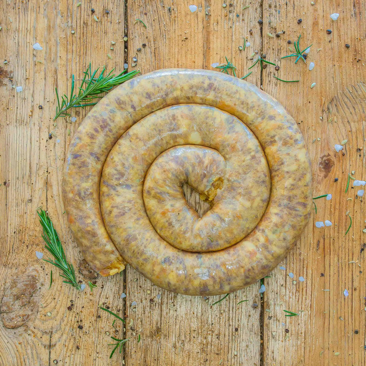 A thick coil of raw peach boerewors sausage placed directly on a textured wooden table, surrounded by scattered coarse salt, spices, and fresh rosemary sprigs.