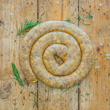 A thick coil of raw peach boerewors sausage placed directly on a textured wooden table, surrounded by scattered coarse salt, spices, and fresh rosemary sprigs.