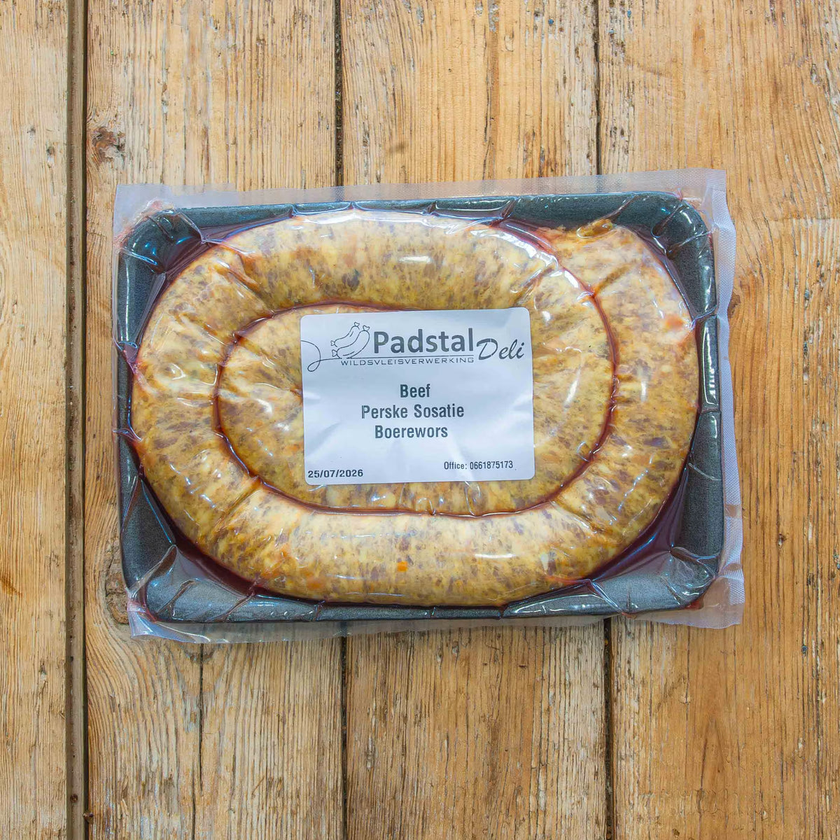 A vacuum-sealed package of raw Padstal Deli Beef Perske Sosatie Boerewors, coiled on a black foam tray and resting on a rustic wooden surface.