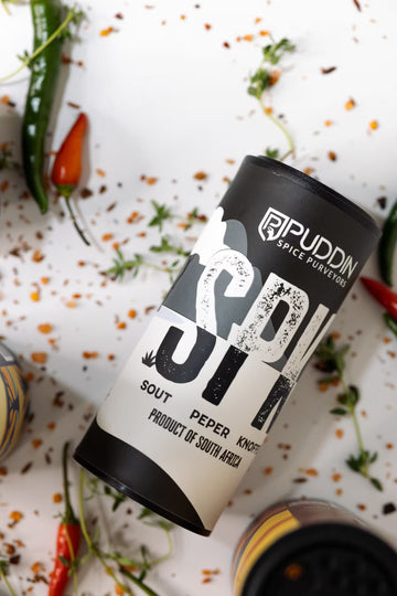 A cylindrical spice jar labeled 'Puddin Spice Purveyors' and 'Sout Pepper Knoffel' lies diagonally on a white surface scattered with chili peppers, herbs, and spices. The jar has a black and off-white label with bold typography.