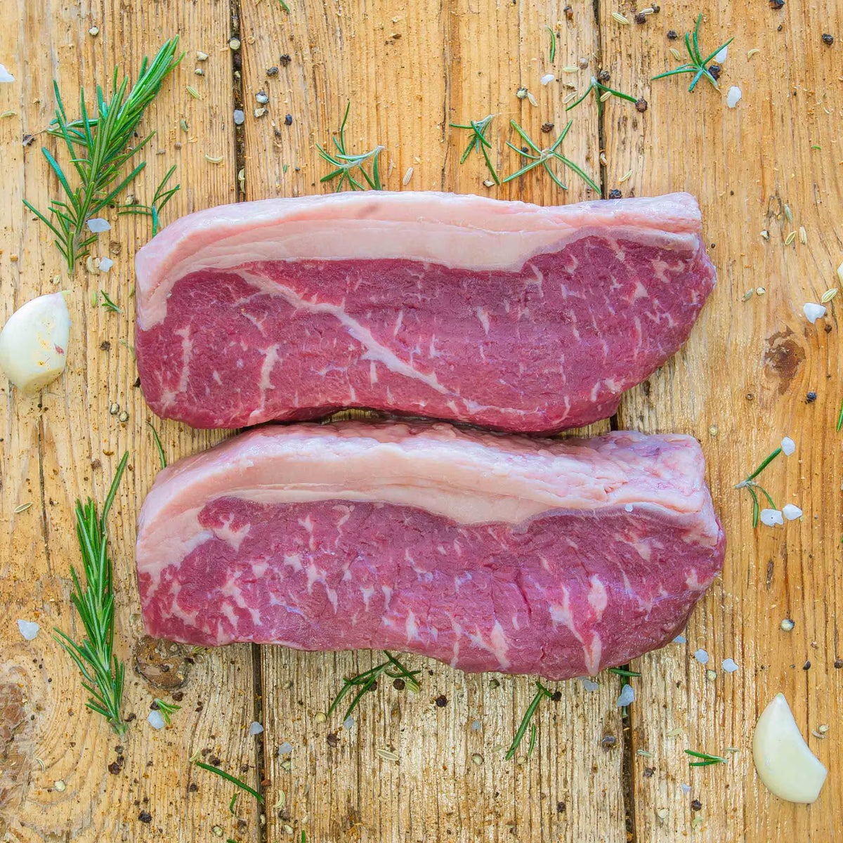 Two raw beef sirloin steaks with thick fat caps resting on a rustic wooden surface, surrounded by scattered fresh rosemary sprigs, garlic cloves, coarse salt crystals, fennel seeds, and black peppercorns.