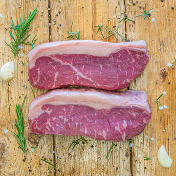 Two raw beef sirloin steaks with thick fat caps resting on a rustic wooden surface, surrounded by scattered fresh rosemary sprigs, garlic cloves, coarse salt crystals, fennel seeds, and black peppercorns.