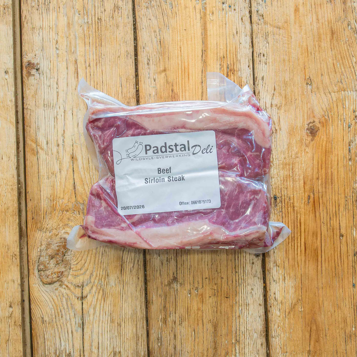 A vacuum-sealed package containing two raw beef sirloin steaks resting on a rustic wooden plank surface. The clear plastic packaging features a white label with the Padstal Deli logo and the text "Beef Sirloin Steak".