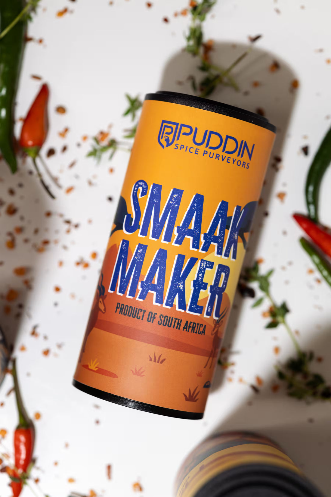 A cylindrical tin of Puddin Spice Purveyors 'Smaak Maker' spice blend, styled on a white surface with scattered spices, chili peppers, and herbs.