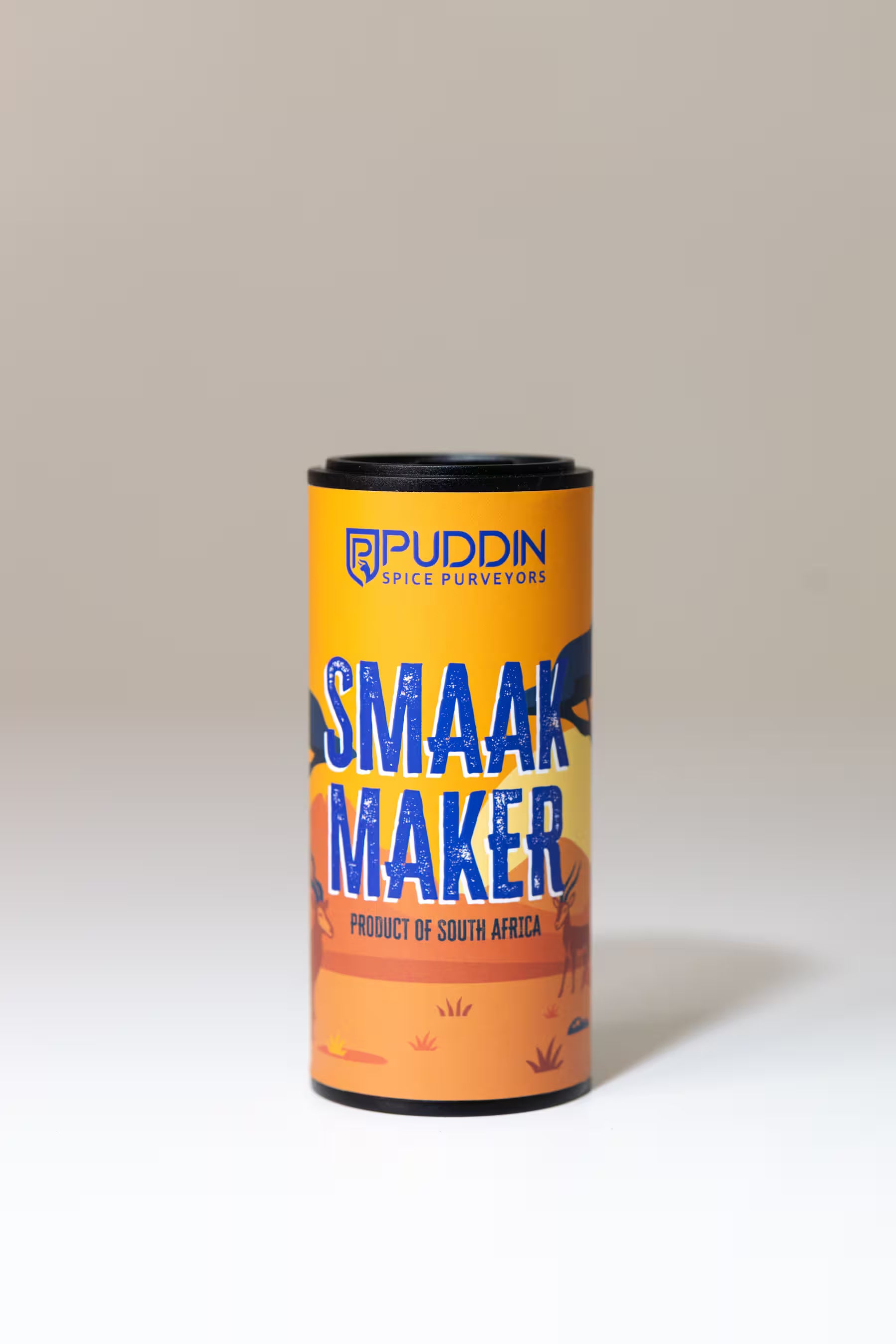 A cylindrical tin of Puddin Smaak Maker, a South African spice mix, set against a neutral beige background. The tin features a bright yellow and orange label with blue text and illustrations of a buck and a sunset.