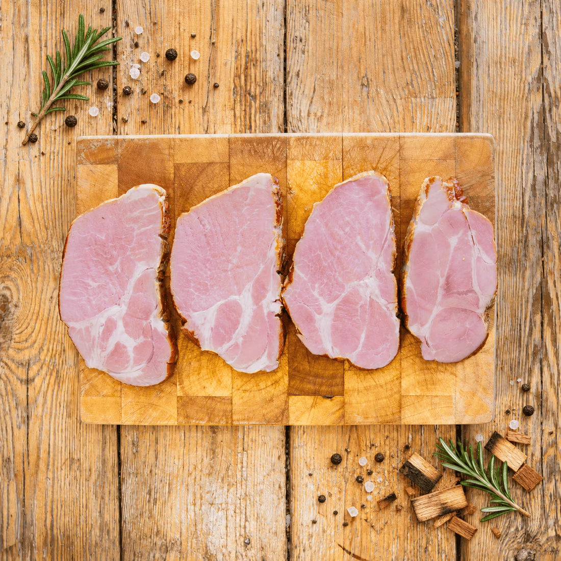 Four individual slices of smoked pork neck arranged side-by-side on a well-used, square wooden chopping board. The board sits on a rustic wooden plank table garnished with fresh rosemary, peppercorns, coarse salt, and wood chips scattered around the edges.
