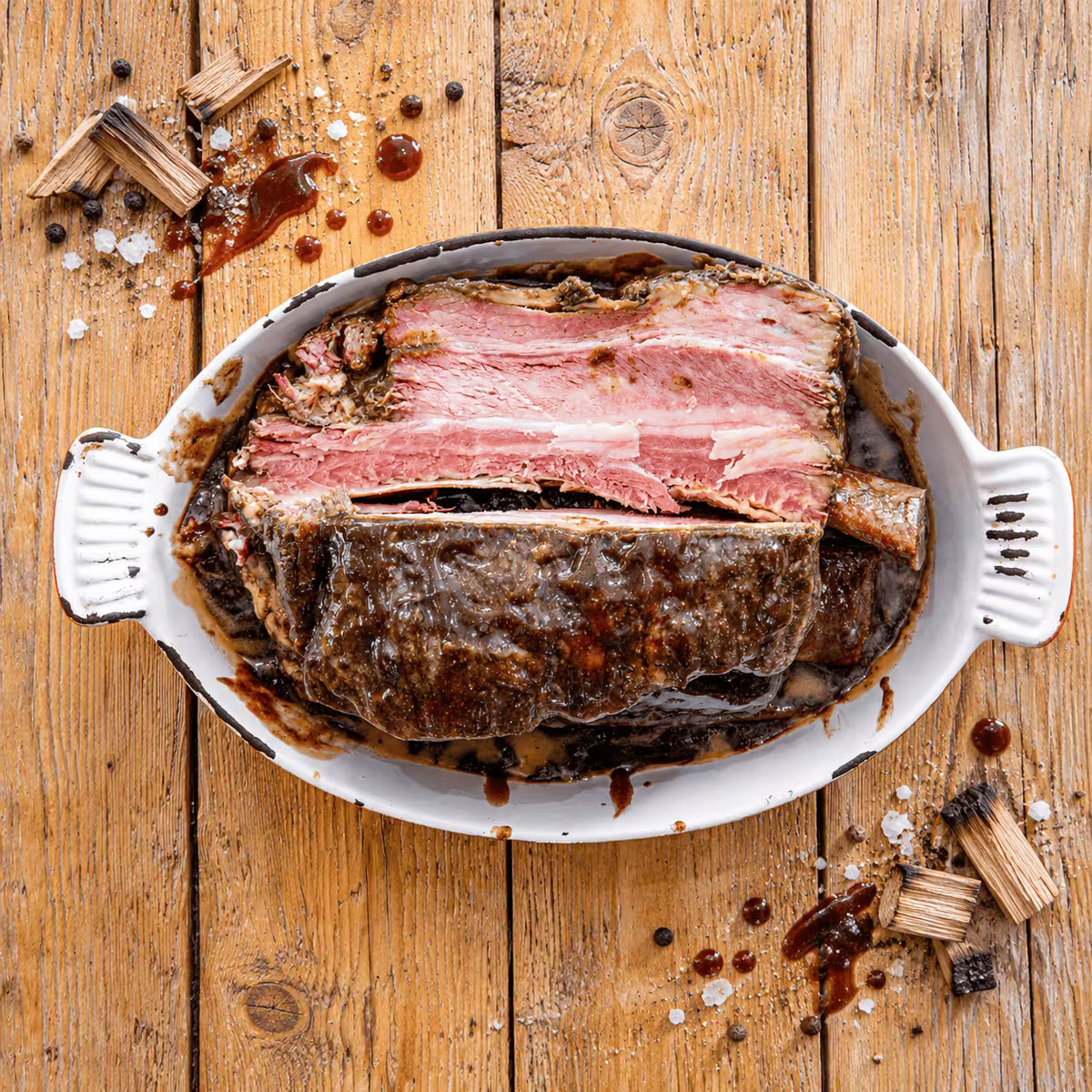 A cooked beef rib heavily glazed in a thick BBQ sauce, presented in a white, rustic oval serving dish on a wooden plank table. The meat is partially sliced, revealing a pink interior with a distinct smoke ring, and a large bone extends from the right side of the cut. Scattered around the serving dish on the wood are drops of BBQ sauce, coarse salt, black peppercorns, and small chunks of charred wood.