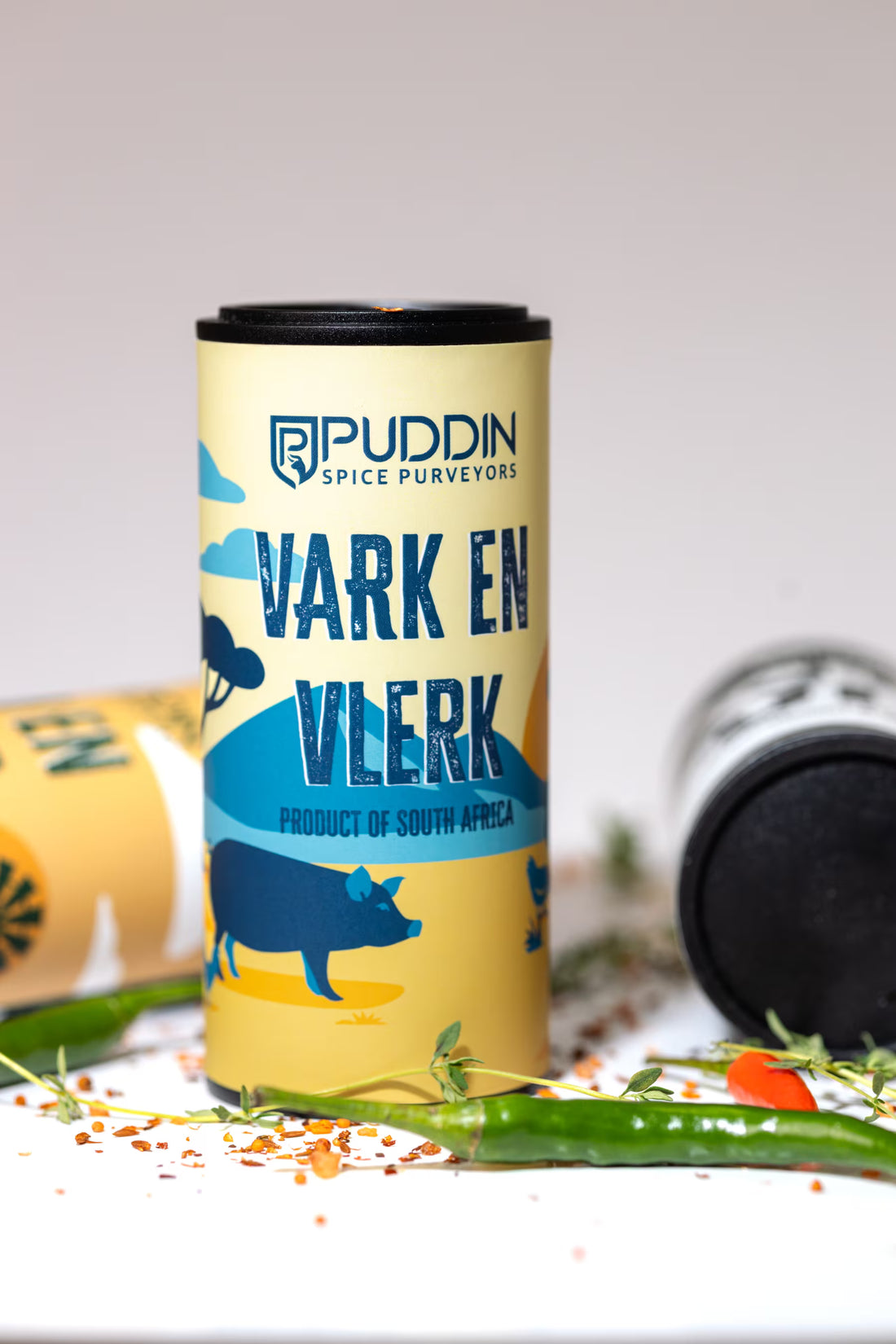 A cylindrical tin of Puddin Spice Purveyors 'Vark En Vlerk' spice mix, styled standing up on a white surface with scattered chili peppers and herbs.