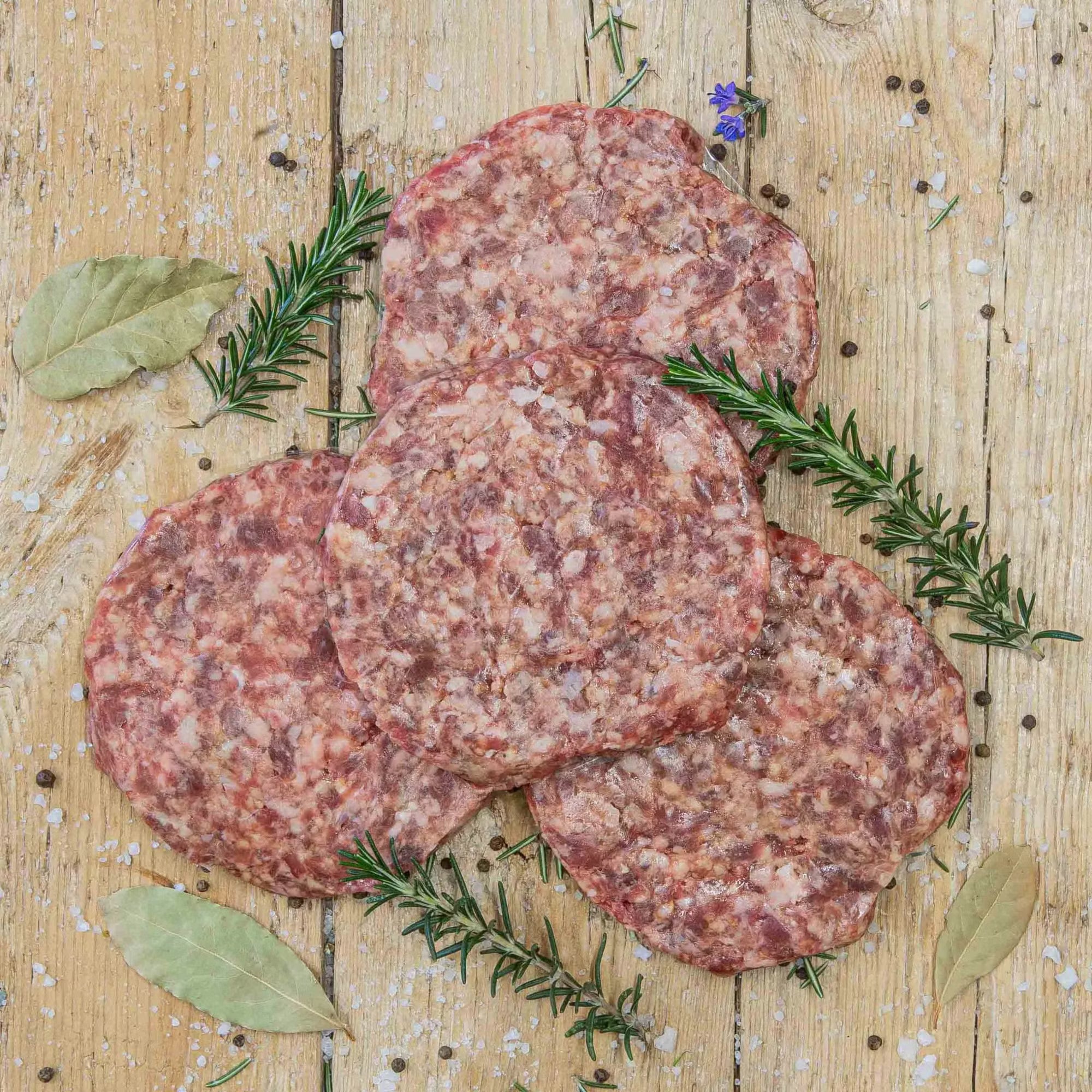 Padstal Deli premium raw burger patties arranged on a rustic wooden background, garnished with dried bay leaves, fresh rosemary sprigs, coarse salt, and peppercorns.