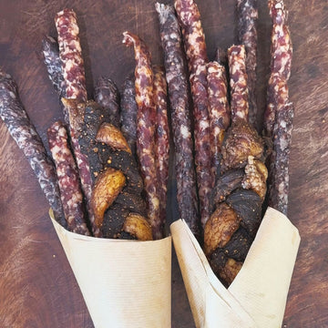 Two Padstal Deli Baby Biltong Bouquets wrapped in light brown craft paper, featuring premium dried meat sticks, droëwors, and braided chilli bites arranged on a dark wooden surface.