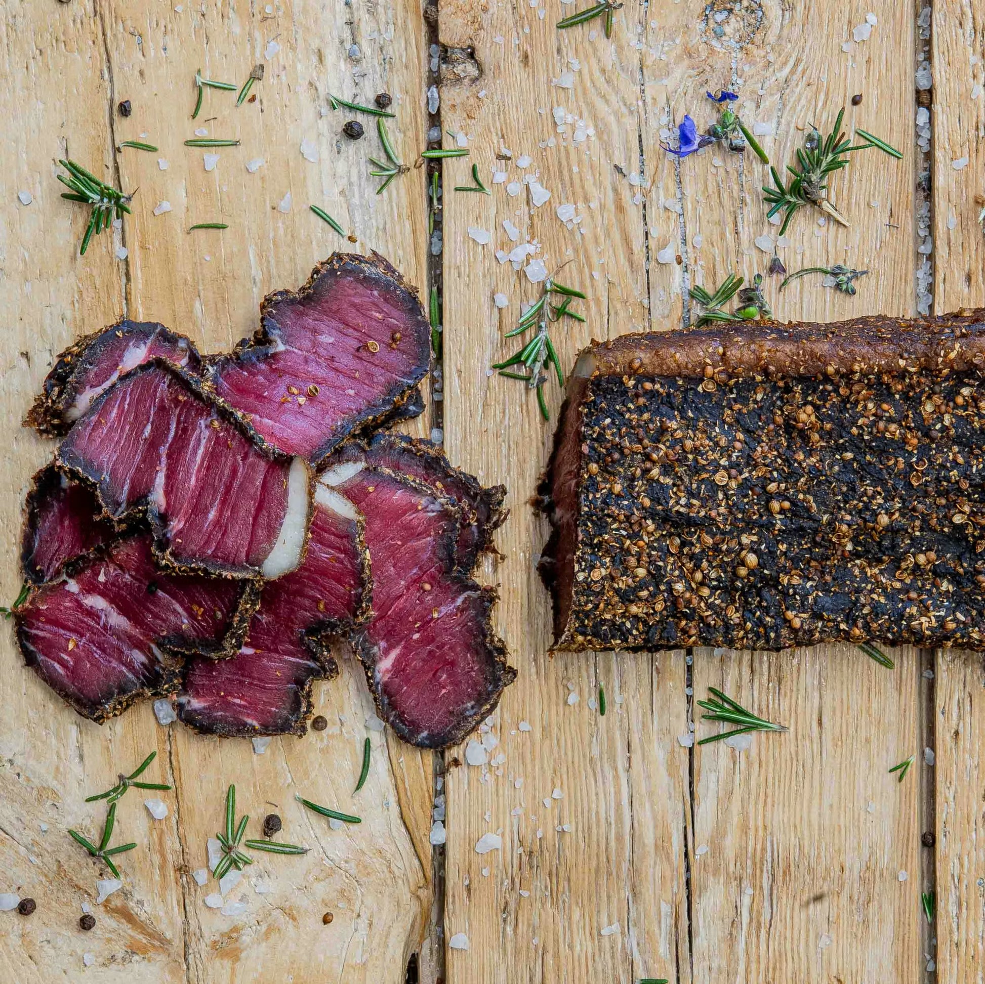 Padstal Deli premium Wet Sliced Beef Biltong pieces with a bright pink center alongside a whole biltong slab on a wooden board, garnished with coarse salt and fresh rosemary.