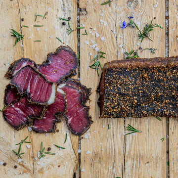 Padstal Deli premium Wet Sliced Beef Biltong pieces with a bright pink center alongside a whole biltong slab on a wooden board, garnished with coarse salt and fresh rosemary.
