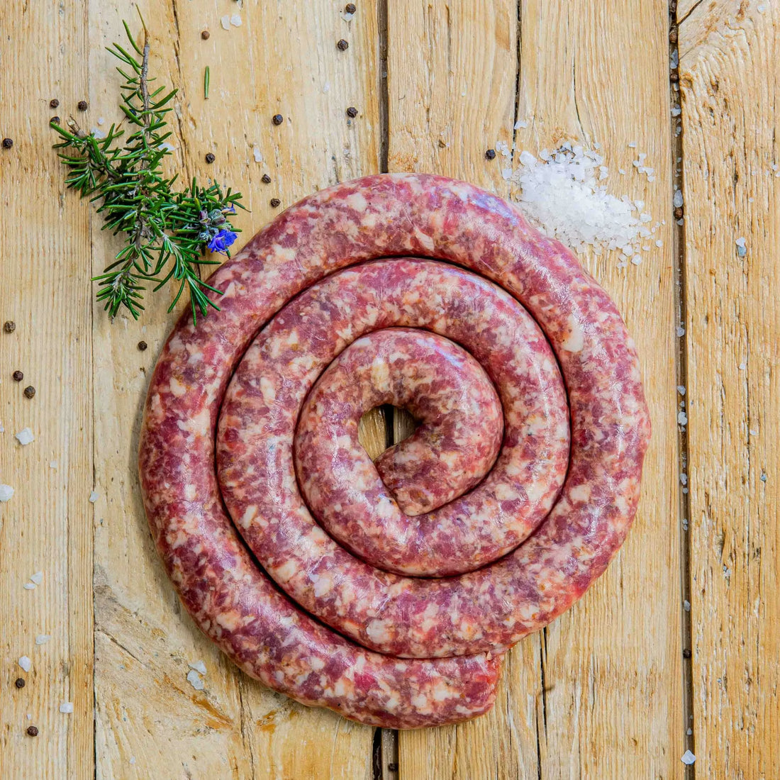 Padstal Deli Artisan Beef Boerewors coiled in a tight spiral on a light rustic wooden background, garnished with coarse sea salt, black peppercorns, and a fresh rosemary sprig.