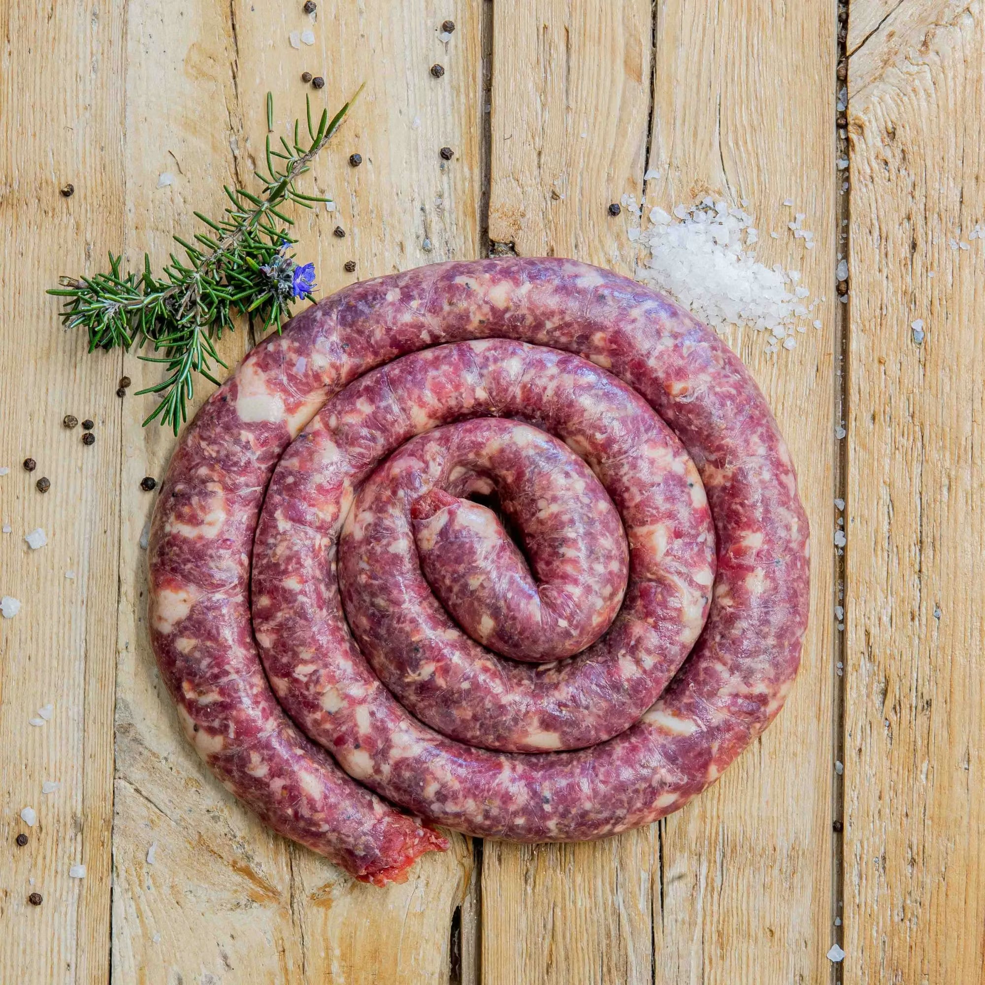Padstal Deli traditional Beef Boerewors coiled in a tight spiral on a light wooden background, garnished with coarse sea salt, black peppercorns, and a sprig of fresh rosemary.