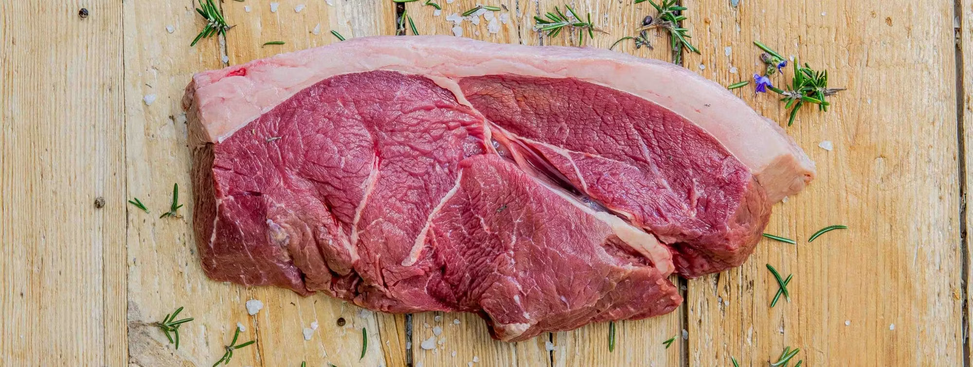 Padstal Deli premium raw Beef Rump Steak showcasing a thick rind of fat, resting on a rustic wooden background styled with coarse salt and fresh rosemary sprigs.