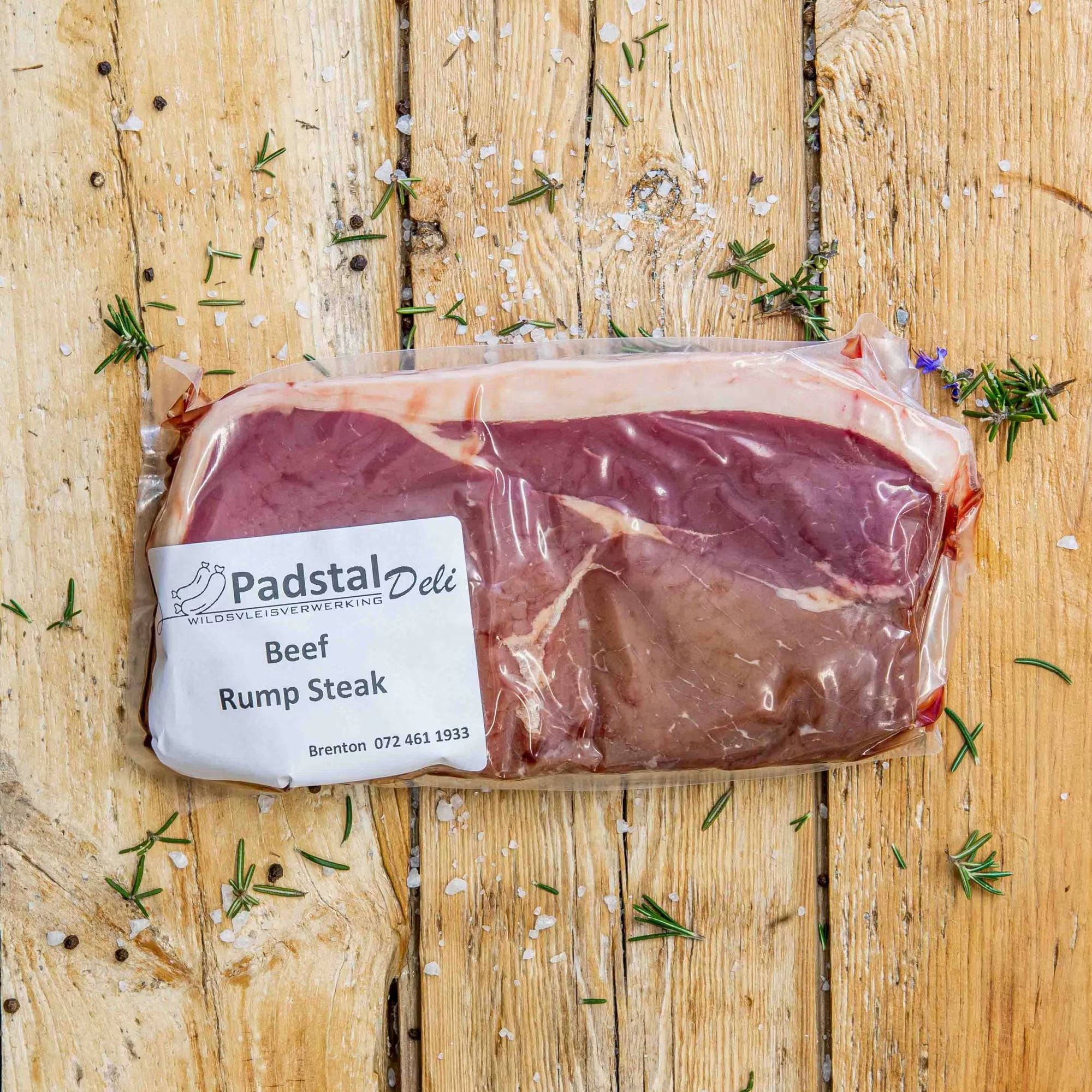 Padstal Deli vacuum-sealed premium Beef Rump Steak cut resting on a rustic wooden background, decorated with scattered coarse salt, peppercorns, and fresh rosemary.