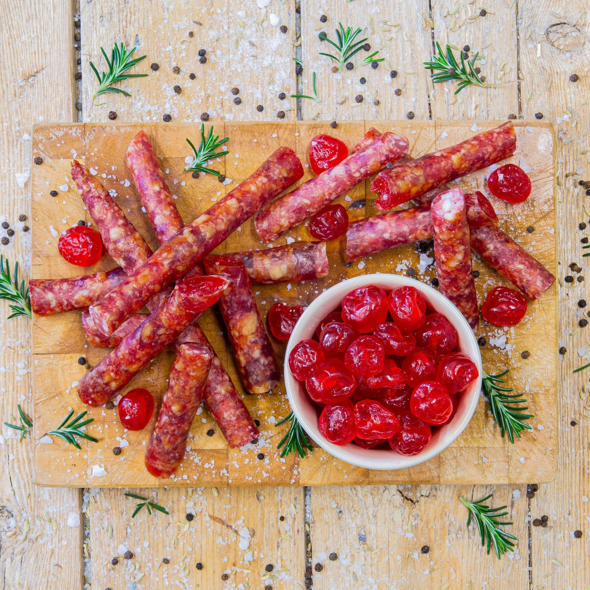 Padstal Deli gourmet Cherry Cabanossi sticks scattered on a wooden cutting board, served with a small bowl of bright red glacé cherries, coarse salt, and rosemary.