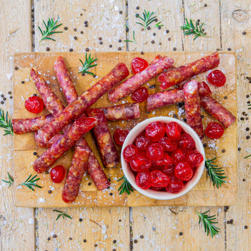 Padstal Deli gourmet Cherry Cabanossi sticks scattered on a wooden cutting board, served with a small bowl of bright red glacé cherries, coarse salt, and rosemary.