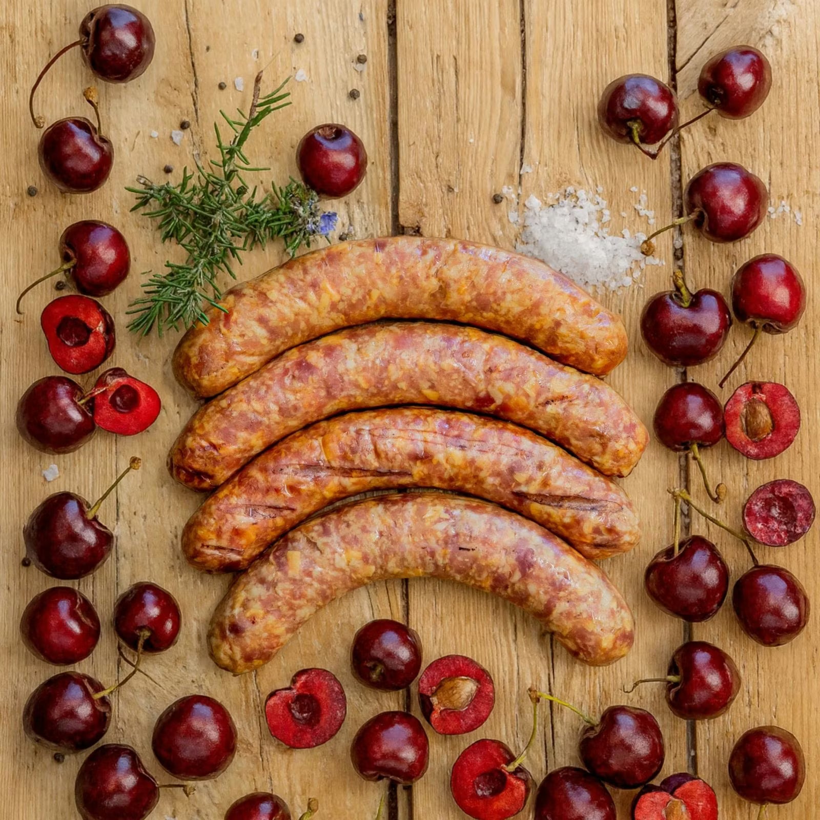Ready-to-eat Cherry Gerookte Kaaswors arranged on a wooden board, surrounded by fresh red cherries, coarse salt, peppercorns, and fresh rosemary.