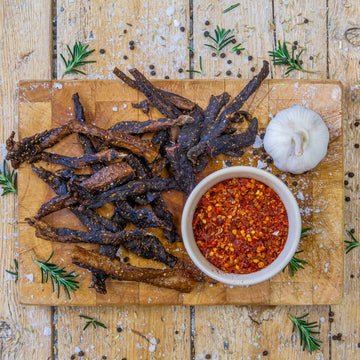 Padstal Deli spicy beef Chilli Bites piled on a wooden cutting board next to a small bowl of crushed dried chili flakes and a whole garlic bulb.