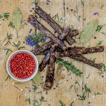 Padstal Deli Pink Pepper Droëwors Stokkies arranged in a rustic flat-lay on a wooden board, served with a small bowl of pink peppercorns, coarse salt, and fresh rosemary.