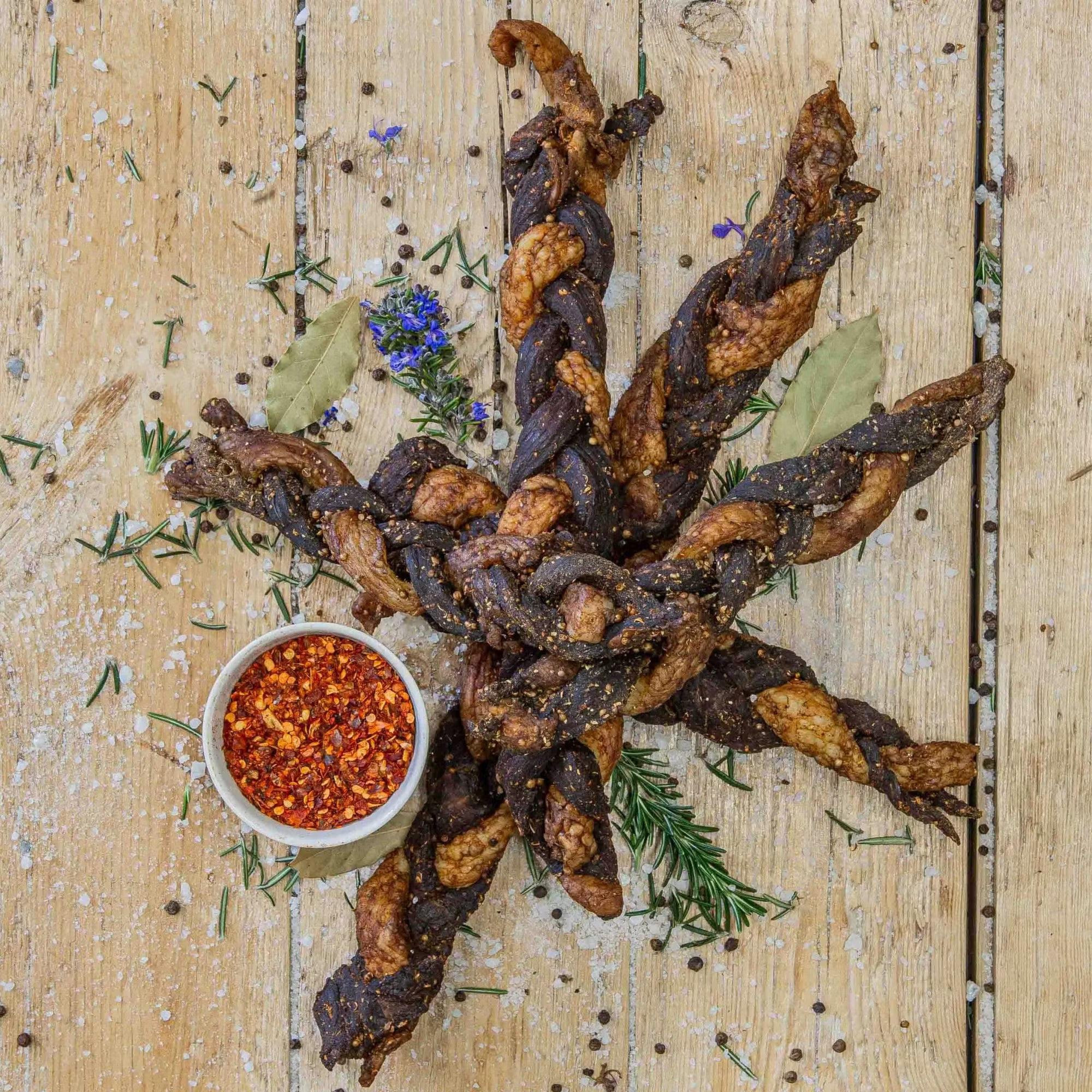 Padstal Deli Twisted Chilli Peri-Peri Chilli Bites arranged in a star pattern on a wooden background, served with a small white bowl of crushed red chili flakes and fresh herbs.