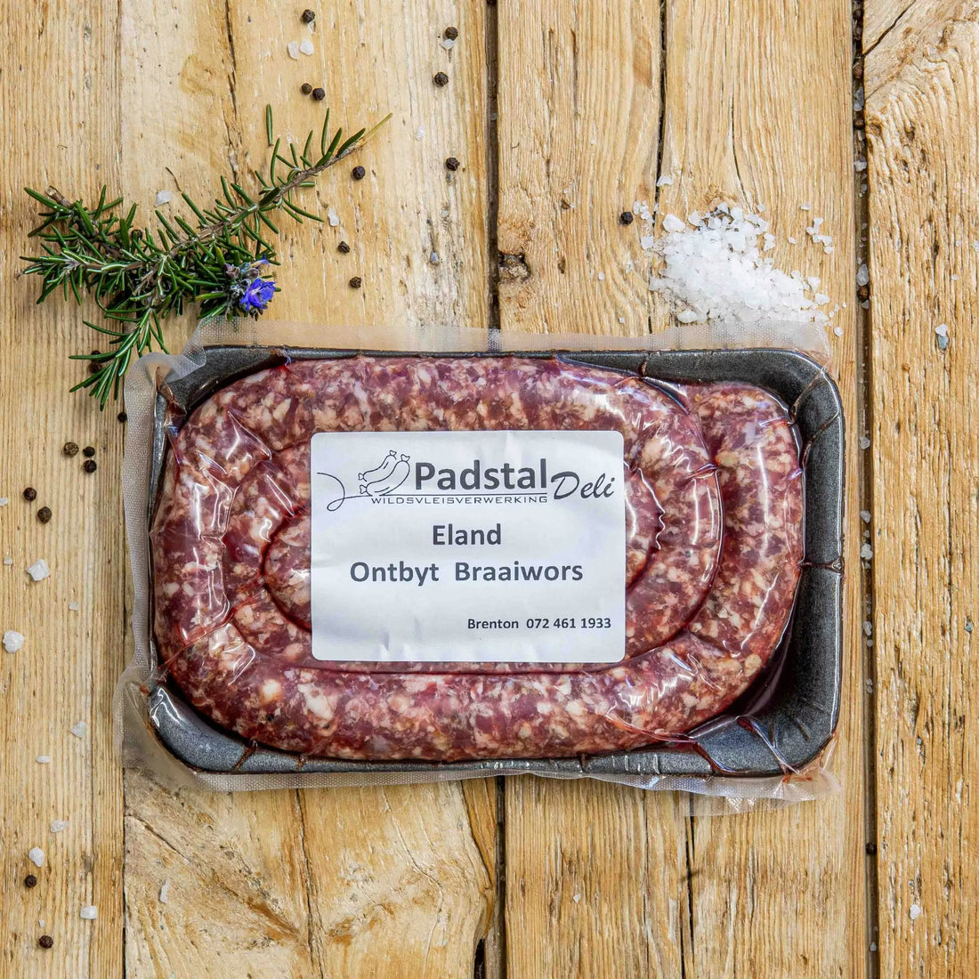 Padstal Deli Ontbyt Braaiwors coiled in a tight spiral on a light wood background, garnished with a pile of coarse sea salt, black peppercorns, and fresh rosemary.